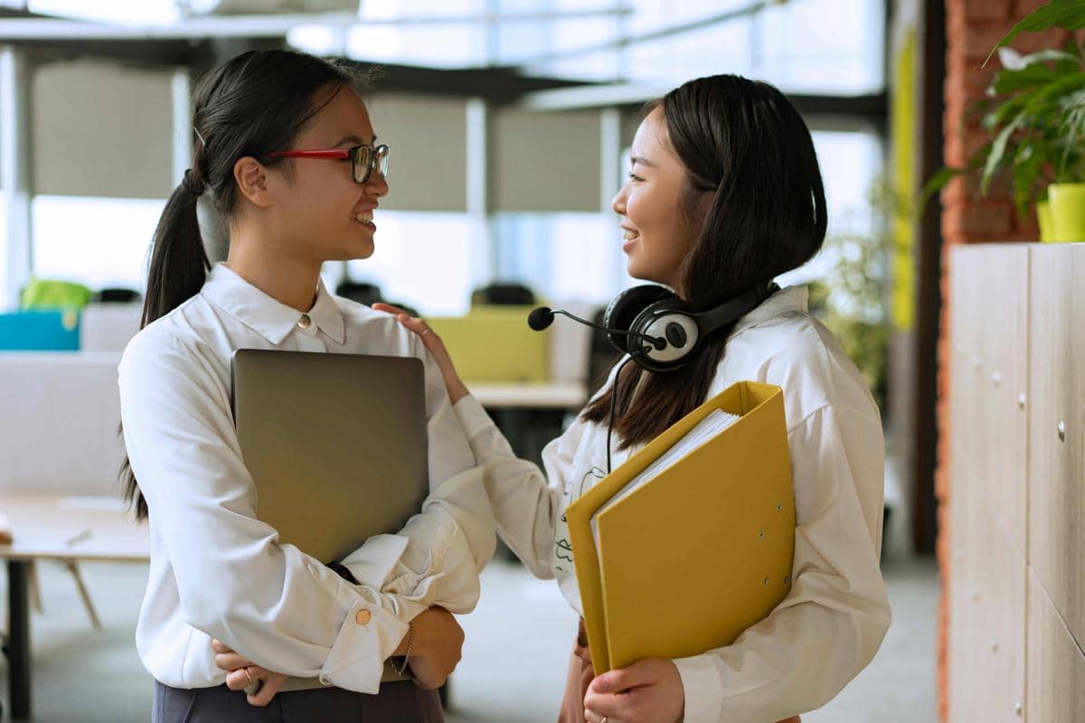 Dua perempuan muda berbicara sambil tersenyum di kantor modern, satu memegang laptop dan lainnya membawa map kuning serta headphone di leher.