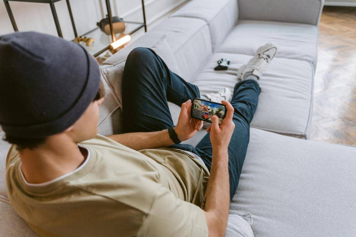 Seorang pria mengenakan beanie dan kaus santai sedang berbaring di sofa sambil bermain game di ponsel pintar.