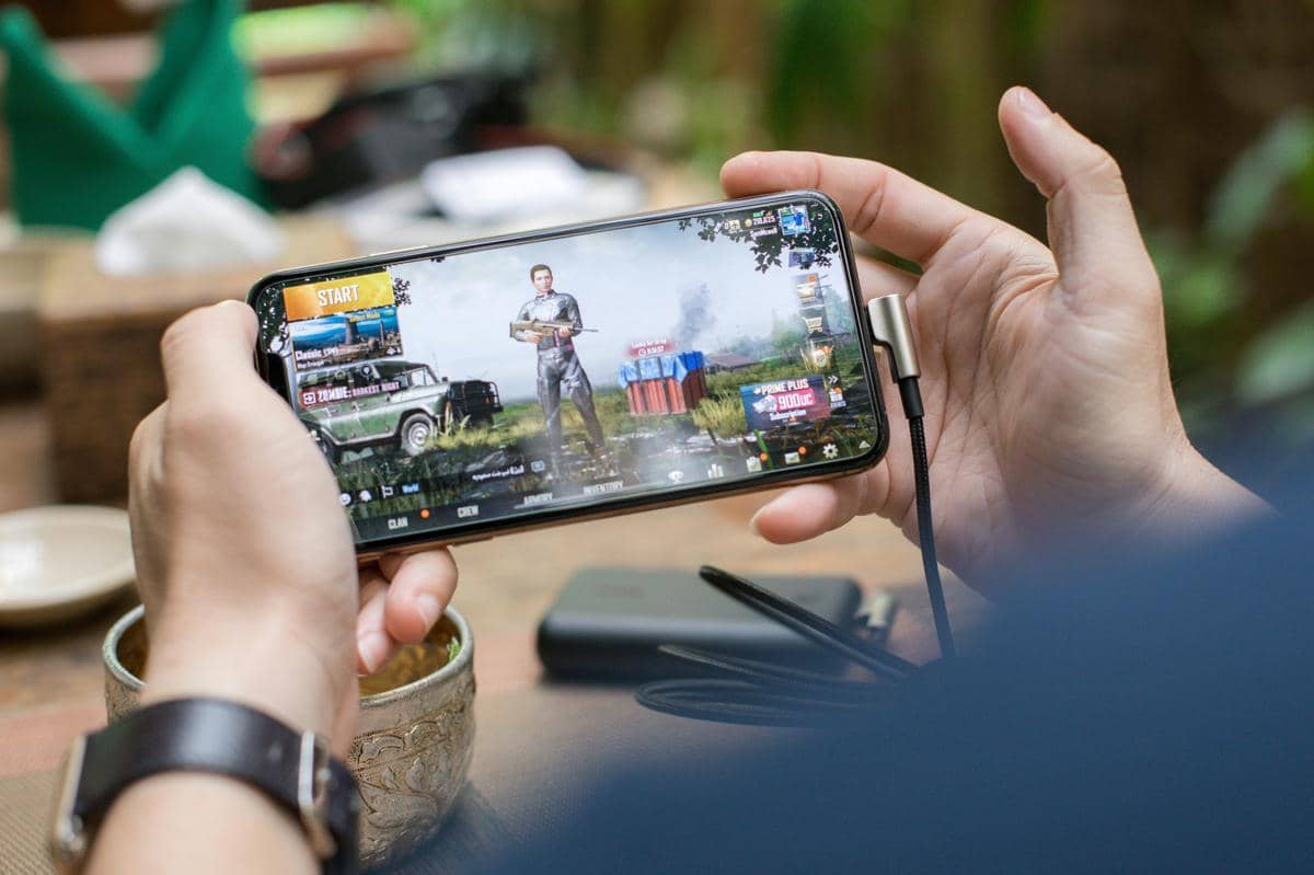 Seseorang memegang ponsel dan memainkan game mobile dengan tampilan karakter serta kendaraan di layar dalam suasana santai.
