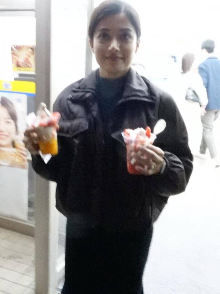 Seorang wanita mengenakan jaket gelap memegang dua gelas es krim dengan topping buah di tangan, berdiri di area luar ruangan yang terang.