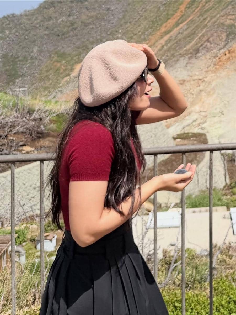 Seorang wanita mengenakan topi baret krem, kacamata hitam, dan kaus merah marun sedang berdiri di luar ruangan di bawah sinar matahari.