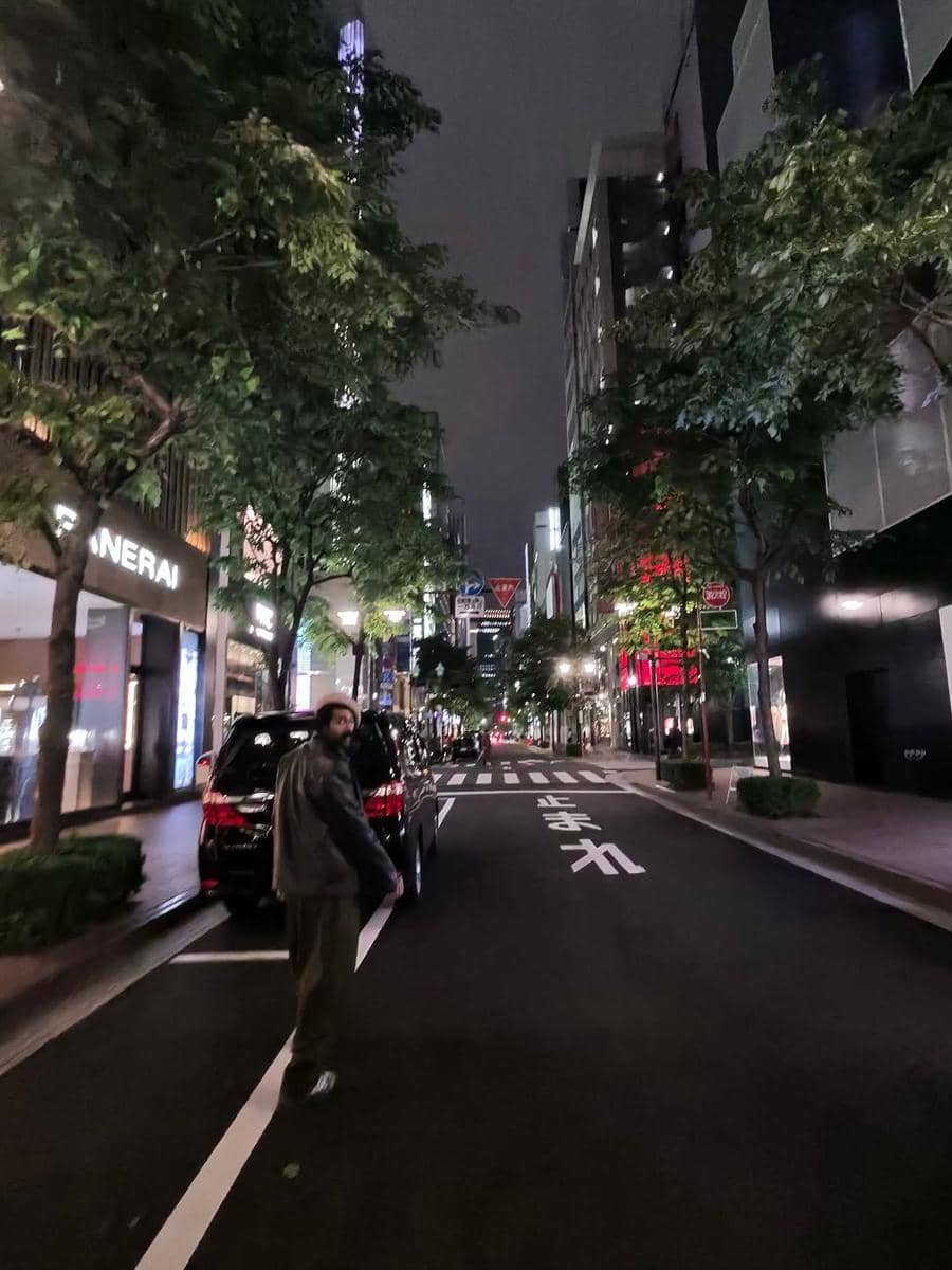 Seorang pria berjalan di jalan kota yang sepi pada malam hari, dikelilingi gedung tinggi dan pepohonan dengan lampu kota yang menyala.