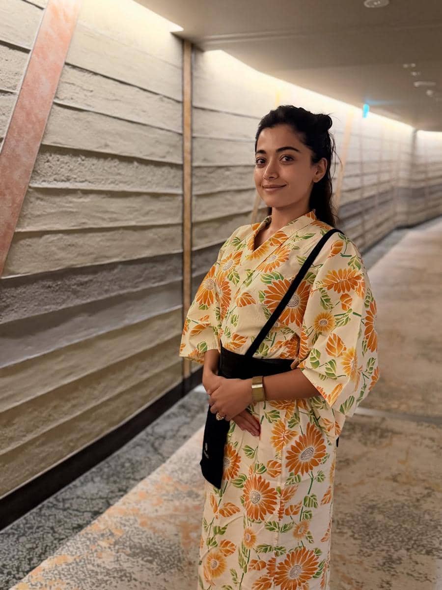 Seorang wanita mengenakan kimono bermotif bunga oranye dan hijau berdiri di koridor dengan dinding bertekstur modern dan pencahayaan hangat.