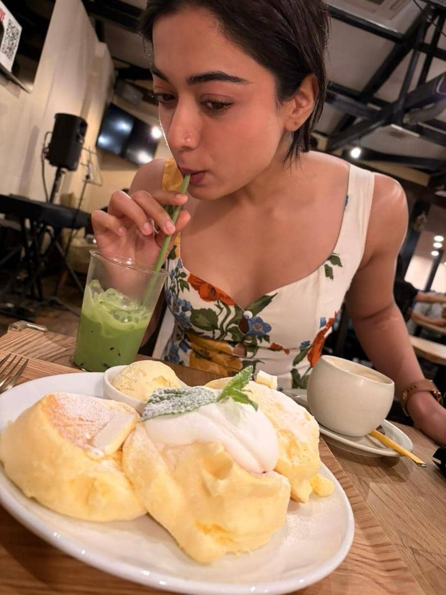 Seorang wanita mengenakan gaun bermotif bunga sedang menikmati minuman hijau di kafe dengan sepiring pancake lembut dan es krim di depannya.