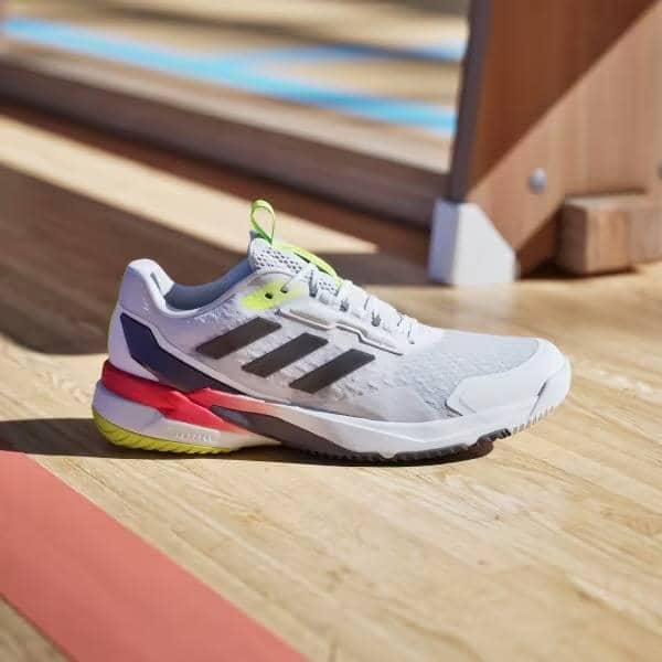 Sepatu olahraga Adidas Crazyflight berwarna putih dengan aksen hitam, merah muda, dan kuning neon di lantai kayu lapangan indoor.