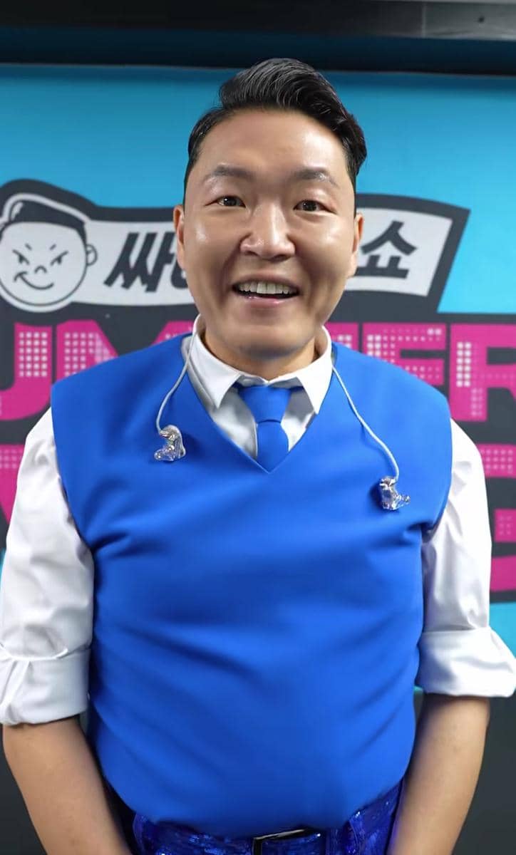 Seorang pria mengenakan rompi biru dan dasi biru dengan kemeja putih, tersenyum di depan latar belakang berwarna biru dan hitam.