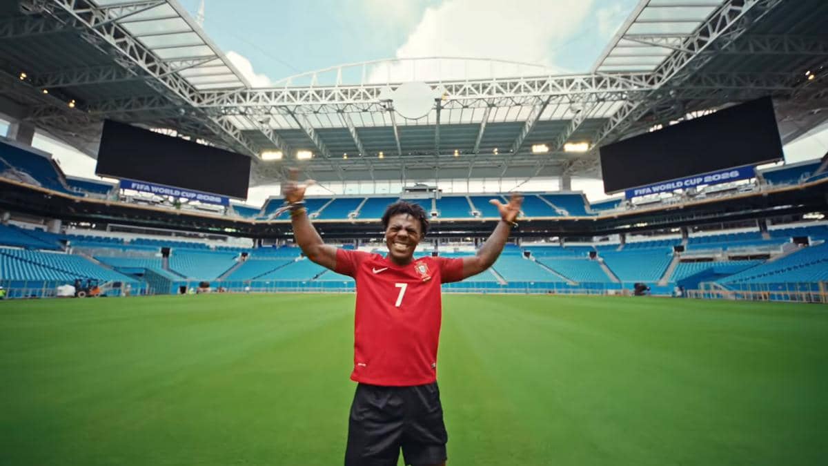 Seorang pria mengenakan jersey merah bernomor 7 berdiri di tengah stadion sepak bola besar dengan tribun biru dan atap terbuka.