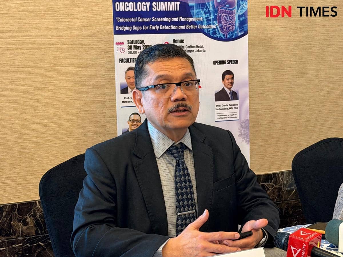 Seorang pria mengenakan jas dan dasi sedang berbicara di depan mikrofon pada acara bertema onkologi dengan latar poster seminar medis.