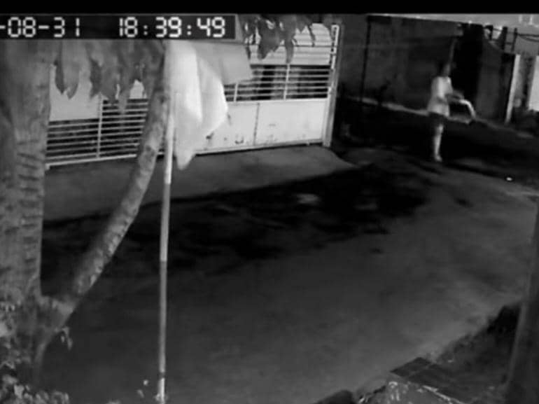 Tangkap layar CCTV memperlihatkan seseorang menyiram air ke jalan di depan rumah pada malam hari dengan gerbang tertutup di latar belakang.