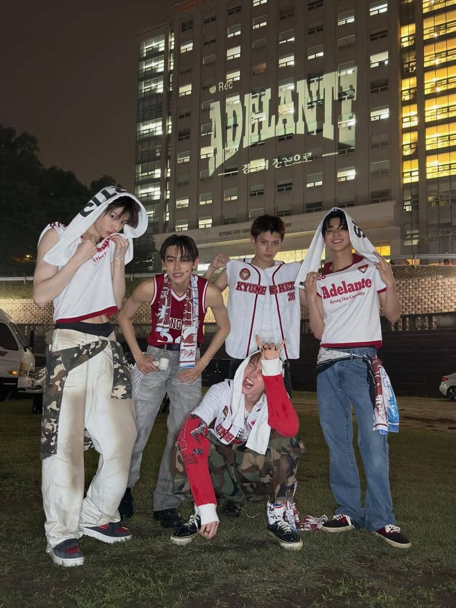 Lima anak muda berpose santai di malam hari di depan gedung bertingkat dengan tulisan besar 'Adelante' yang diproyeksikan di dinding.