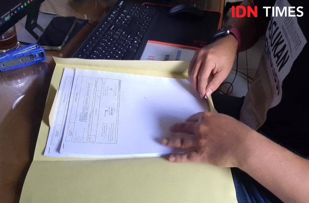 Seseorang menyiapkan berkas laporan dalam map cokelat di atas meja kerja dengan komputer dan alat tulis di sekitarnya.