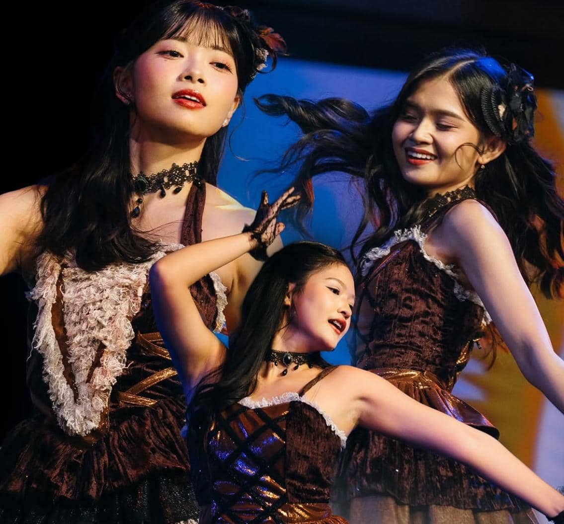 Anggota grup idola JKT48 tampil di atas panggung dengan kostum cokelat berhias renda dan ekspresi penuh semangat saat menari.