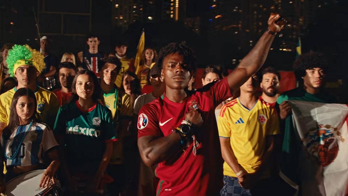 Seorang pria mengenakan jersey Portugal mengangkat tangan dengan latar belakang sekelompok orang memakai kostum tim sepak bola dari berbagai negara.