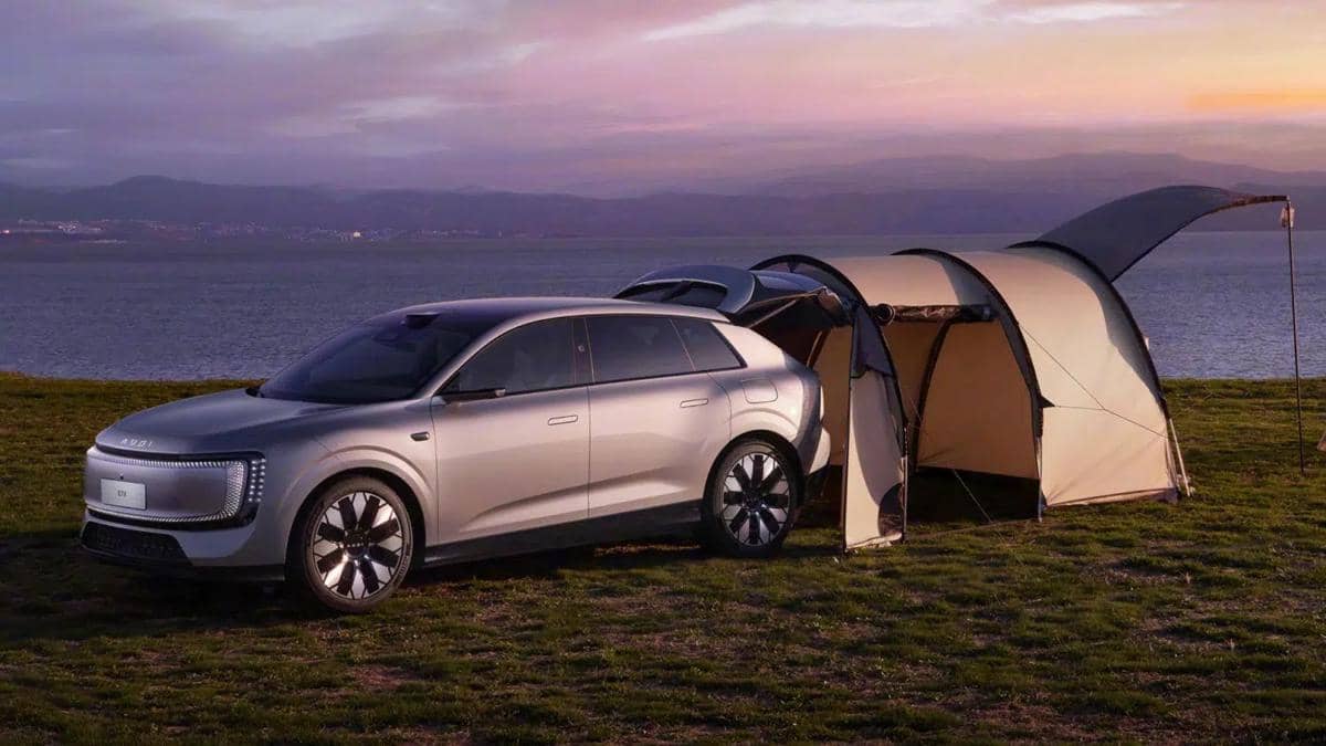 Mobil listrik Audi E7X berwarna perak diparkir di tepi danau dengan tenda terpasang di bagian belakang saat matahari terbenam.