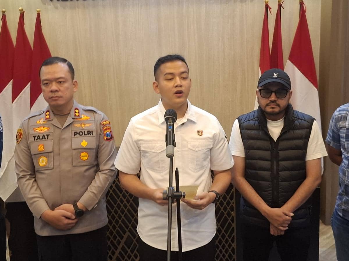 Seorang perwira polisi berdiri di depan mikrofon bersama dua pria lainnya di ruangan dengan latar bendera merah putih.