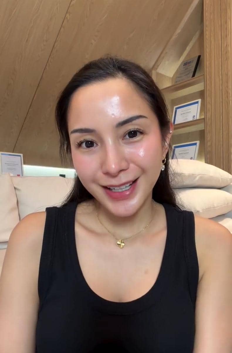 Seorang perempuan berambut panjang mengenakan tank top hitam tersenyum di ruangan dengan dinding kayu dan beberapa sertifikat di rak belakang.
