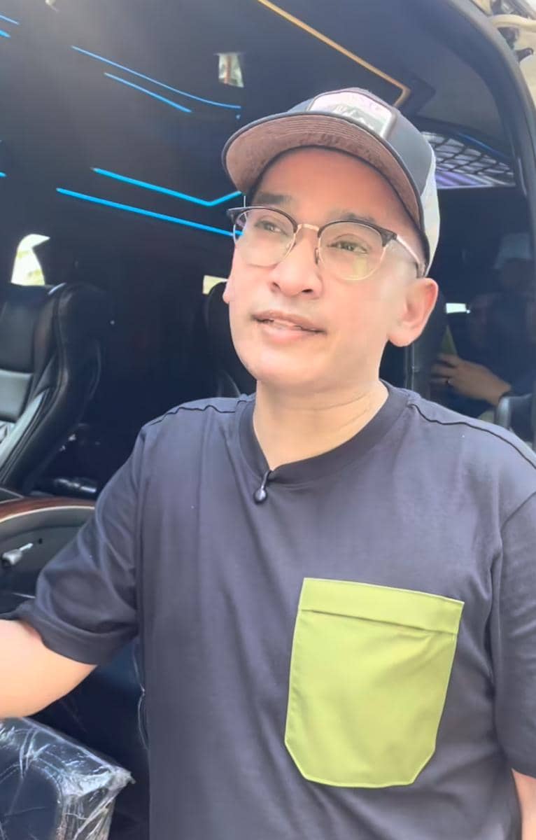 Seorang pria mengenakan kacamata, topi, dan kaus hitam dengan saku hijau sedang berdiri di dekat mobil dengan interior mewah.