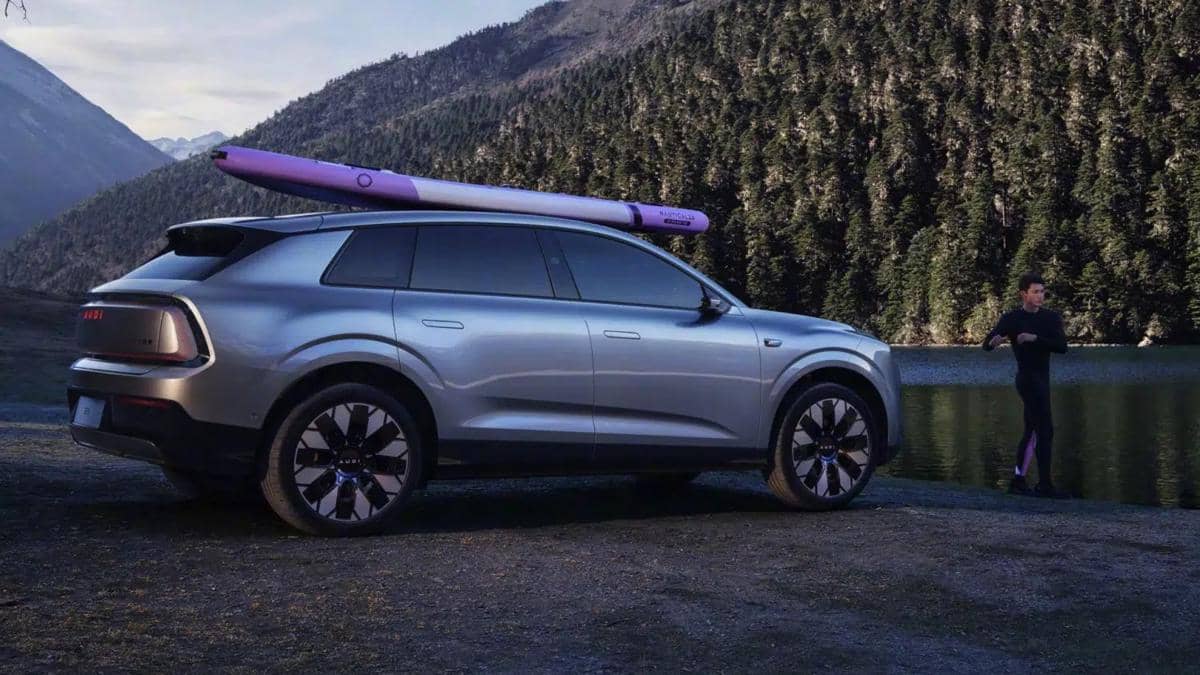 SUV Audi E7X berwarna perak dengan papan selancar di atapnya diparkir di tepi danau berlatar pegunungan dan pepohonan hijau.