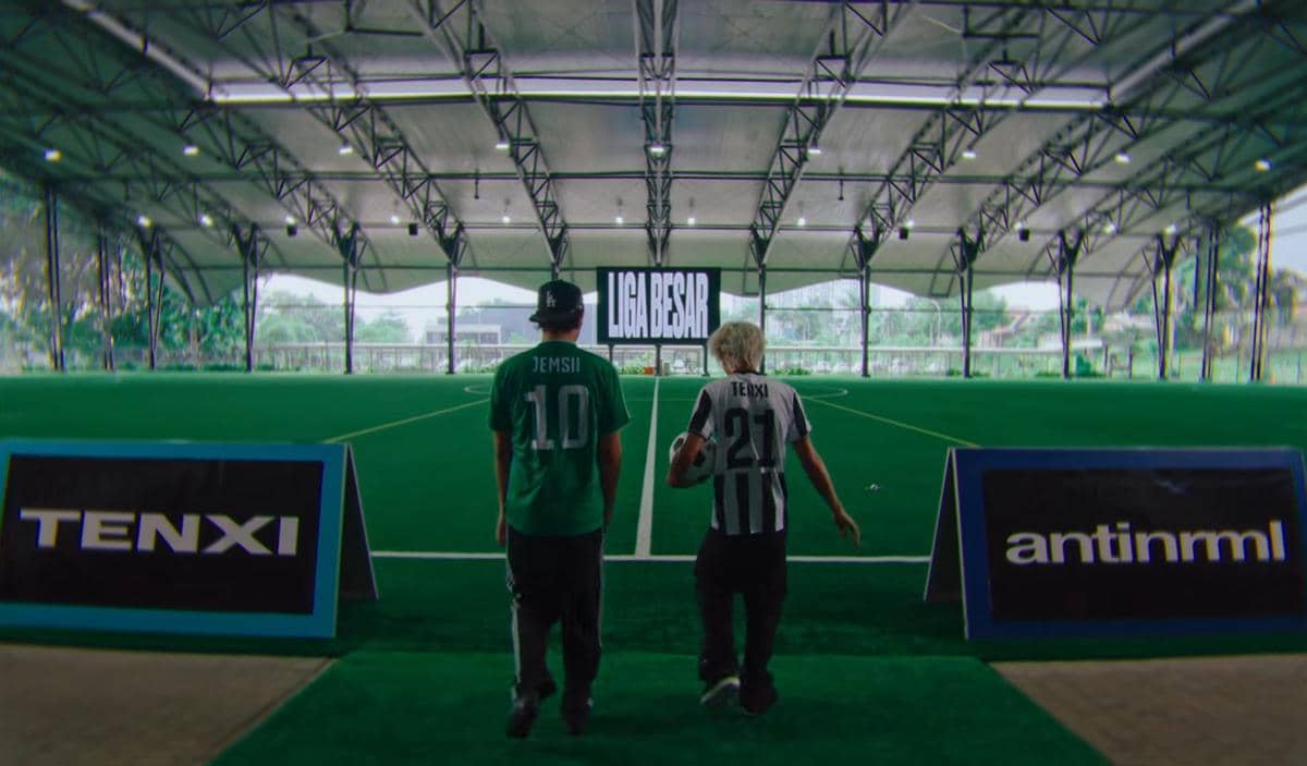 Dua orang mengenakan jersey bertuliskan Tenxi dan Jemsii berjalan menuju lapangan sepak bola indoor dengan layar bertuliskan 'Liga Besar'.