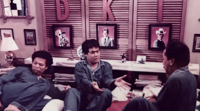Tiga pria mengenakan piyama biru sedang berbincang di kamar dengan latar huruf DKI dan foto-foto di dinding belakang mereka.