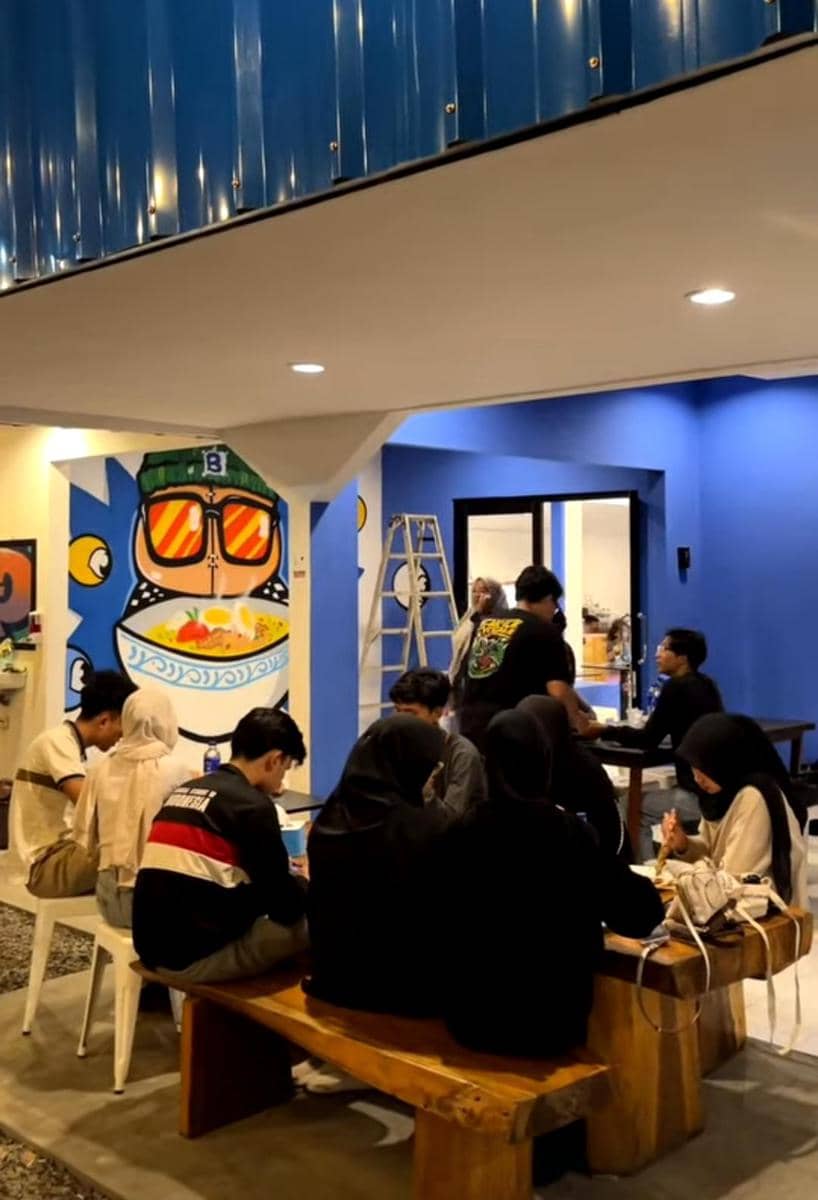 Beberapa orang duduk di meja kayu di Bahagia Canteen dengan dinding biru dan mural mangkuk ramen berwarna cerah di belakang mereka.