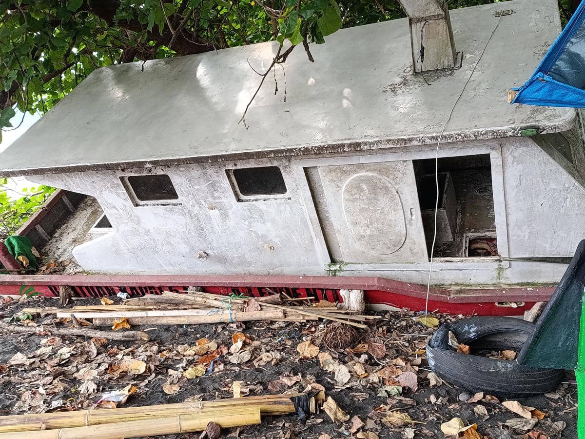 Kapal nelayan berwarna abu-abu dan merah tampak rusak dan tergeletak di tepi pantai dengan daun kering berserakan di sekitarnya.