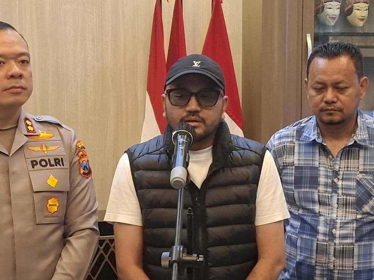 Seorang pria berbicara di depan mikrofon didampingi seorang polisi berseragam dan pria lain di ruangan dengan bendera Indonesia di belakangnya.