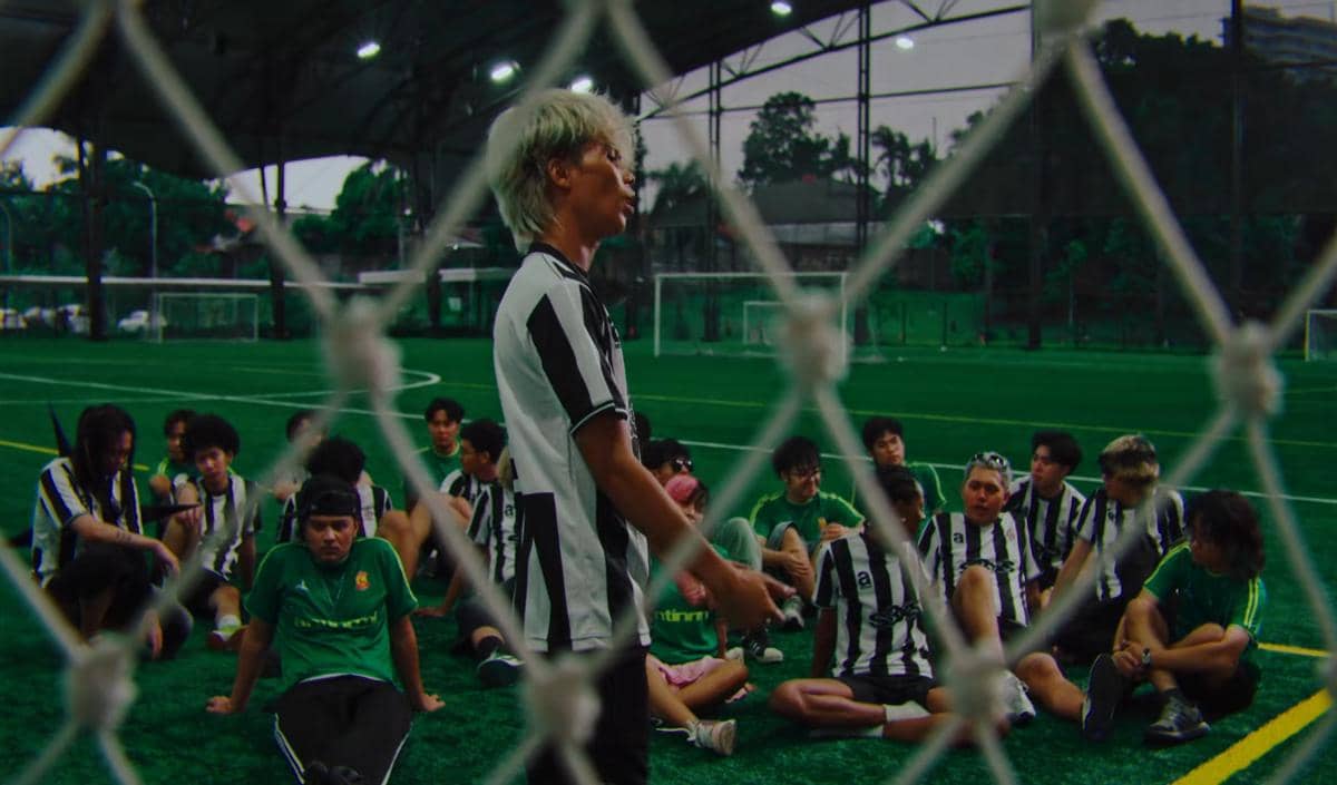Seorang pria berambut pirang mengenakan seragam hitam putih berdiri di lapangan sepak bola indoor di depan sekelompok orang yang duduk.