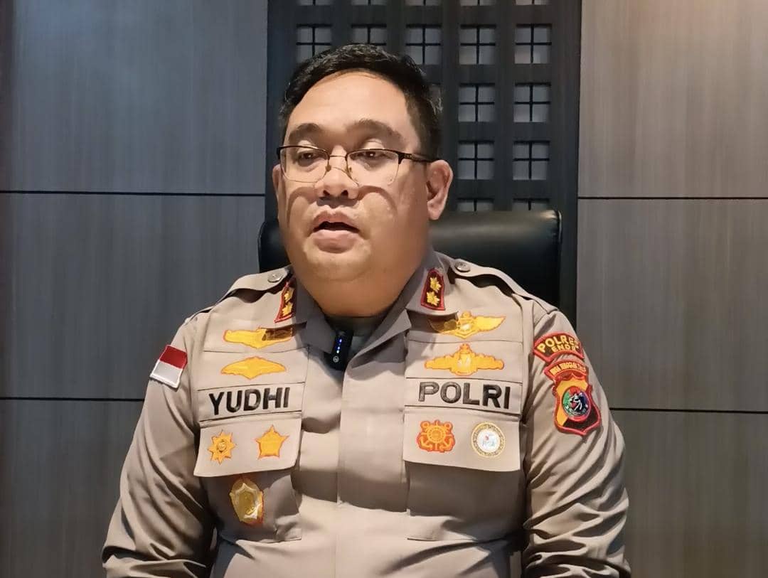 Seorang perwira polisi mengenakan seragam lengkap dengan tanda pangkat dan nama di dada, duduk di kursi di dalam ruangan kantor.