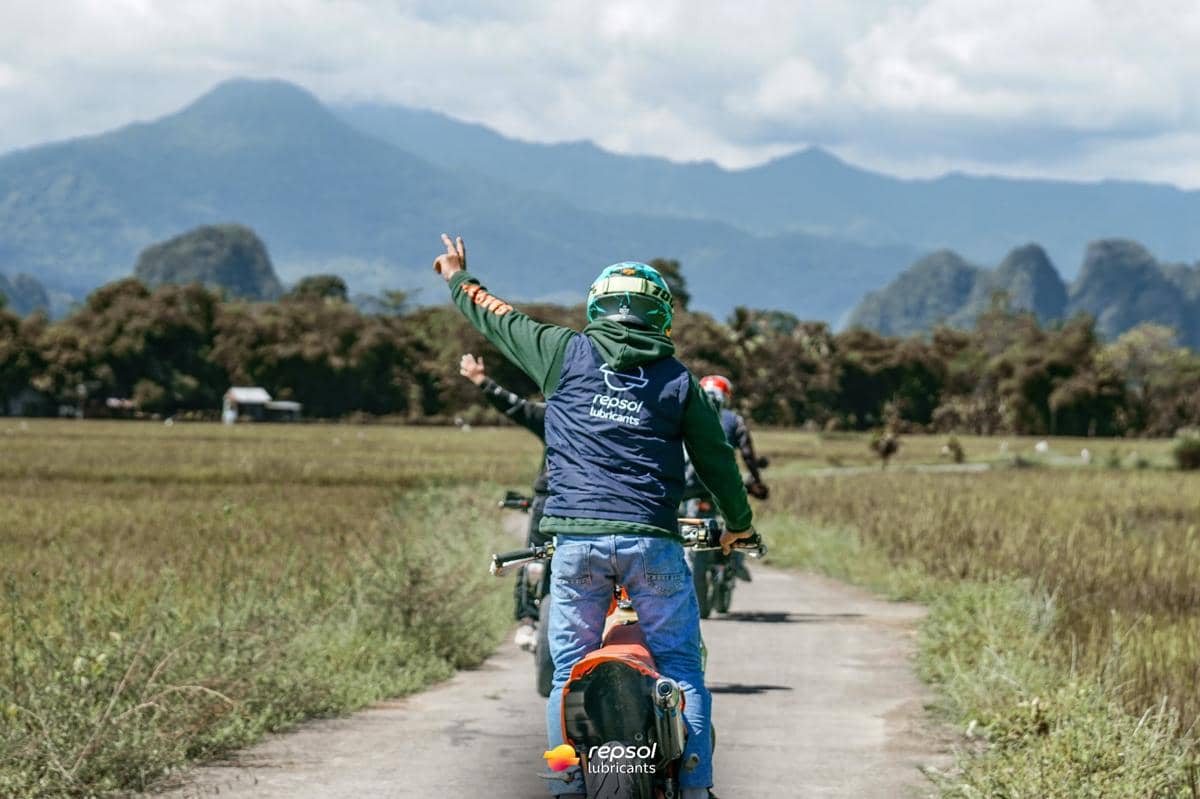 Seorang pengendara motor mengenakan helm dan rompi Repsol mengacungkan tangan di jalan pedesaan dengan latar pegunungan hijau.