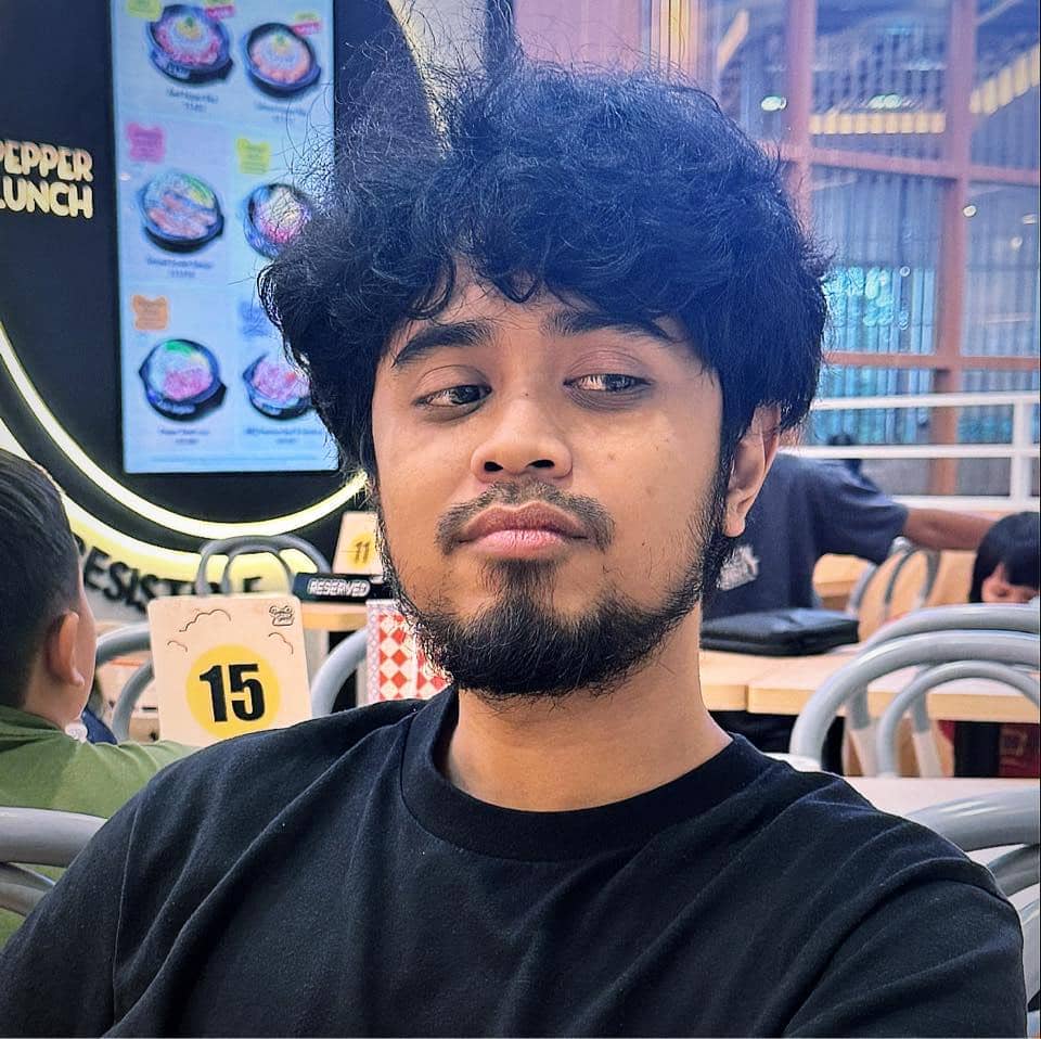 Seorang pria berambut ikal dan berjanggut mengenakan kaus hitam duduk di restoran dengan latar papan menu dan meja bernomor 15.