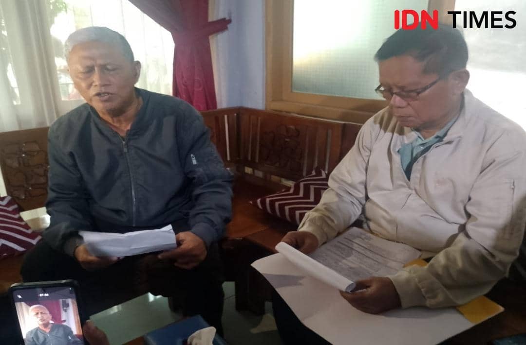 Dua pria lanjut usia duduk di ruang tamu sambil memegang berkas dan surat, tampak serius membahas dokumen pinjaman.