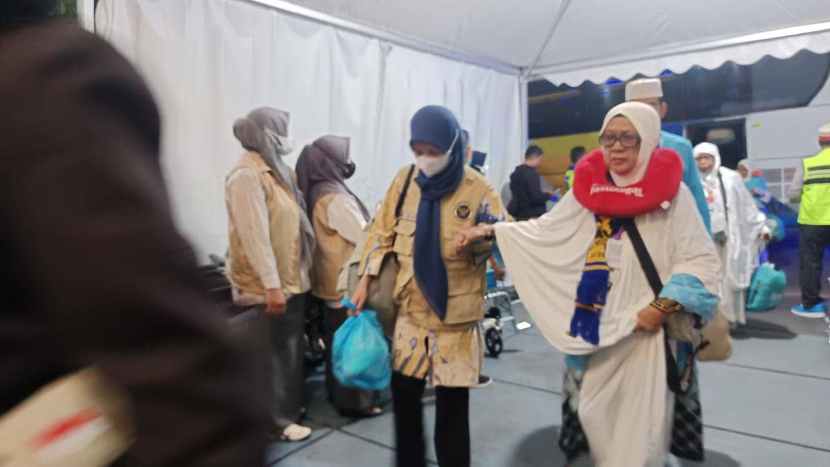 Seorang jemaah haji tiba di Asrama Haji Sudiang Makassar pada malam hari, dibantu petugas yang mengenakan rompi dan hijab.