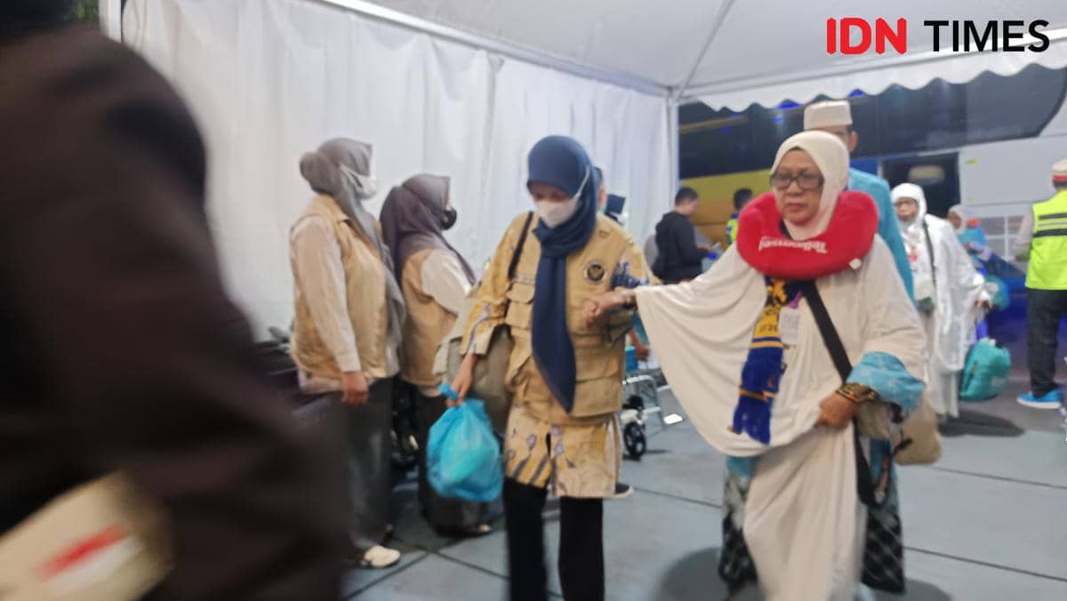 Seorang jemaah haji Kloter 1 Debarkasi Makassar tiba di Asrama Haji Sudiang pada malam hari, didampingi petugas yang mengenakan rompi dan hijab.