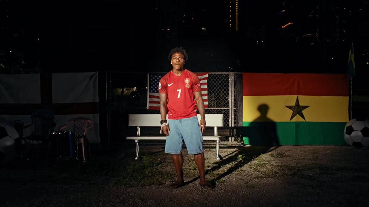 Seorang pria mengenakan jersey merah berdiri di depan bangku taman dengan latar bendera Amerika Serikat dan Ghana pada malam hari.
