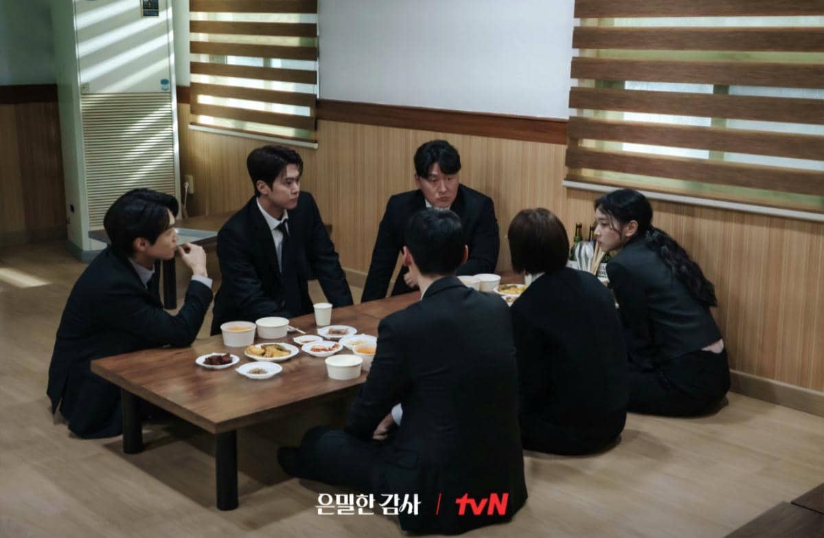 Enam orang berpakaian formal duduk di lantai sekitar meja rendah dengan hidangan Korea di atasnya dalam adegan drama Filing for Love.