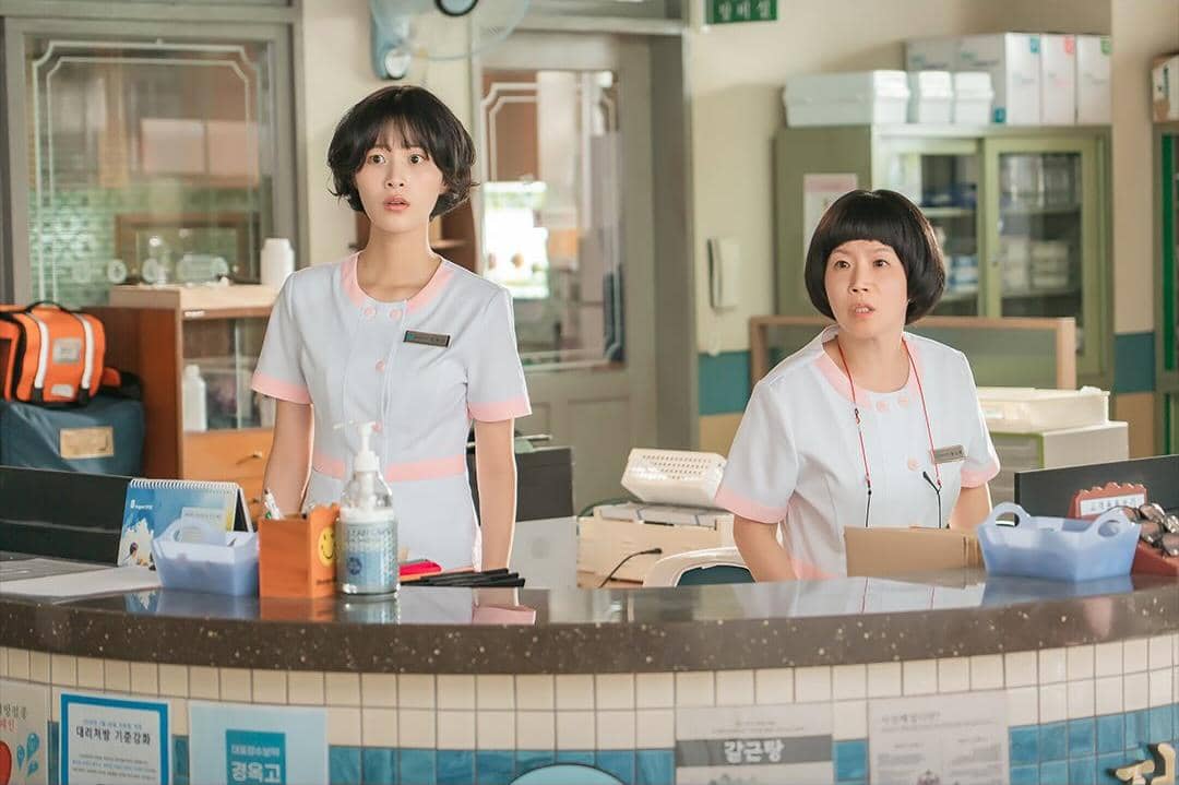 Dua perawat berdiri di meja resepsionis rumah sakit dalam adegan drama Korea Doctor On The Edge, mengenakan seragam putih muda dengan aksen merah muda.