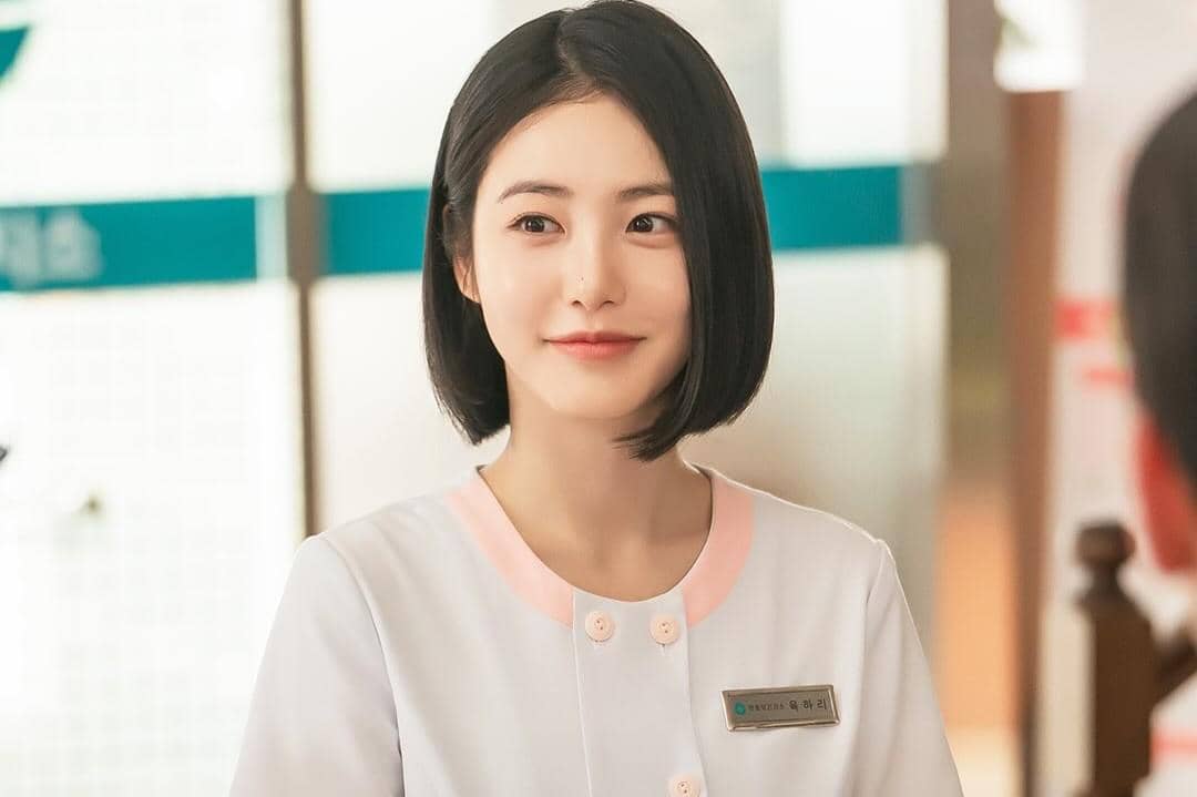 Seorang perempuan berambut pendek mengenakan seragam medis putih dengan kerah merah muda tersenyum di dalam ruangan terang.