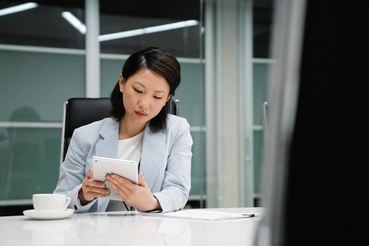 Seorang perempuan mengenakan blazer biru muda sedang menggunakan tablet di meja kantor dengan secangkir kopi di depannya.