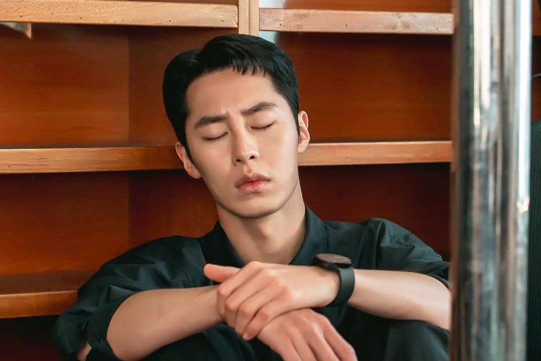 Seorang pria mengenakan kemeja hitam duduk bersandar di rak kayu dengan mata terpejam dan ekspresi lelah di adegan drama Doctor On The Edge.