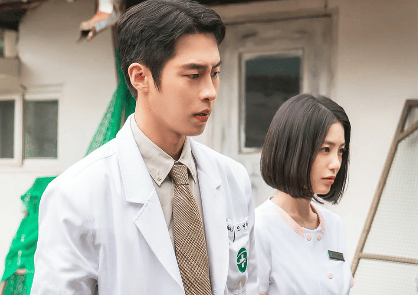 Dua tenaga medis mengenakan jas putih dan seragam perawat sedang berdiri di luar gedung dalam cuplikan drama Korea Doctor On The Edge.