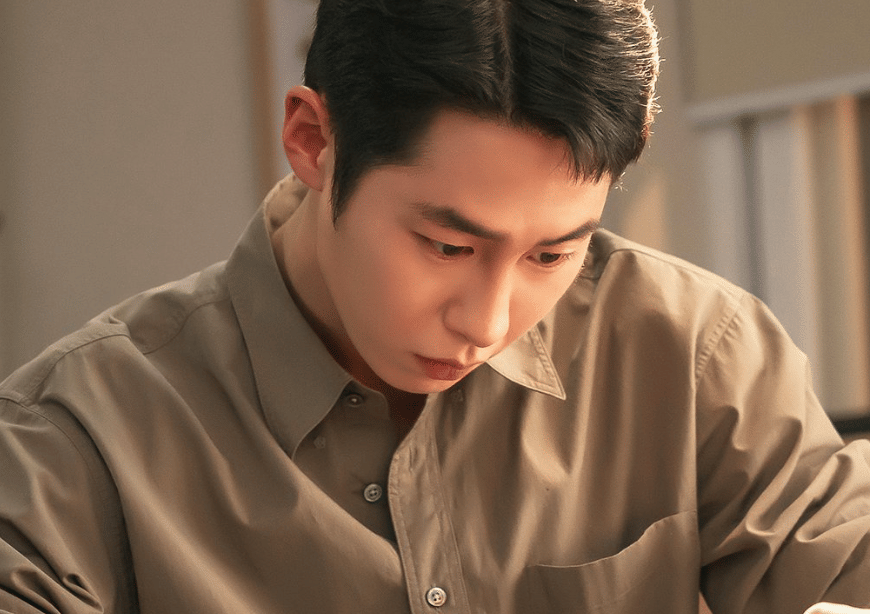 Seorang pria mengenakan kemeja cokelat muda tampak serius menatap ke bawah dalam cuplikan drama Korea Doctor On The Edge.
