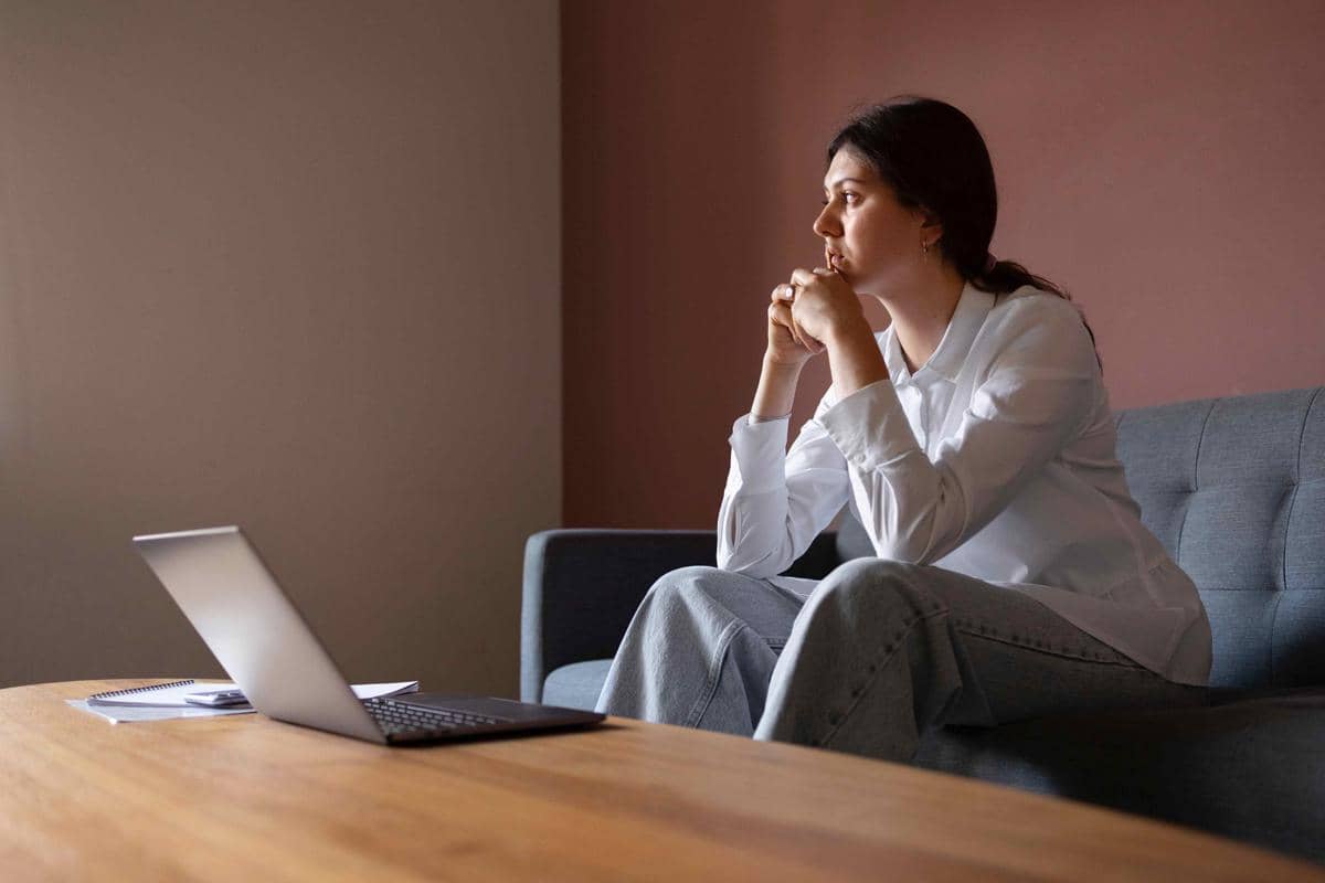 Seorang wanita duduk di sofa dengan ekspresi tenang sambil menatap ke arah jendela, di depannya terdapat laptop di atas meja kayu.
