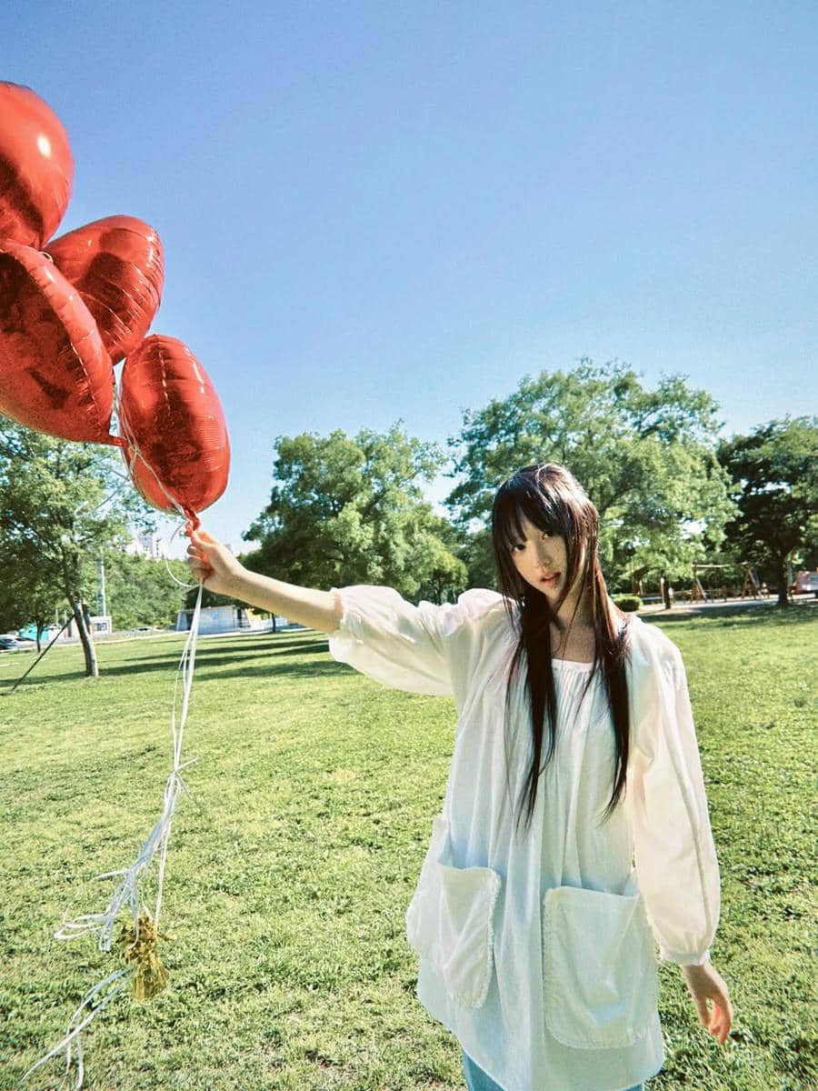 Seorang perempuan berambut panjang mengenakan blus putih memegang balon merah berbentuk hati di taman saat cuaca cerah.