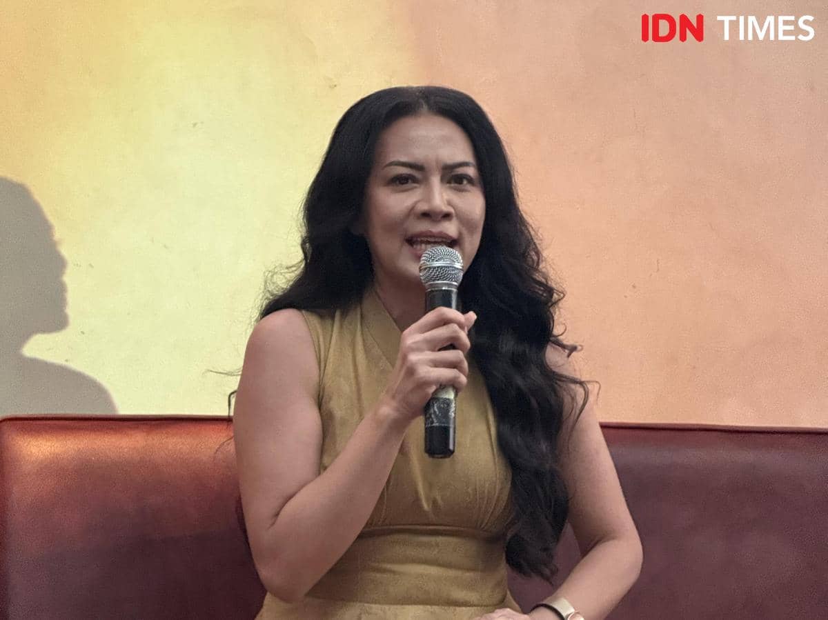 Seorang perempuan berambut panjang mengenakan gaun cokelat muda sedang berbicara sambil memegang mikrofon di acara dalam ruangan.