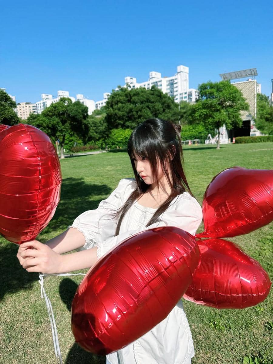 Seorang perempuan berambut panjang mengenakan gaun putih memegang balon merah berbentuk hati di taman saat cuaca cerah.