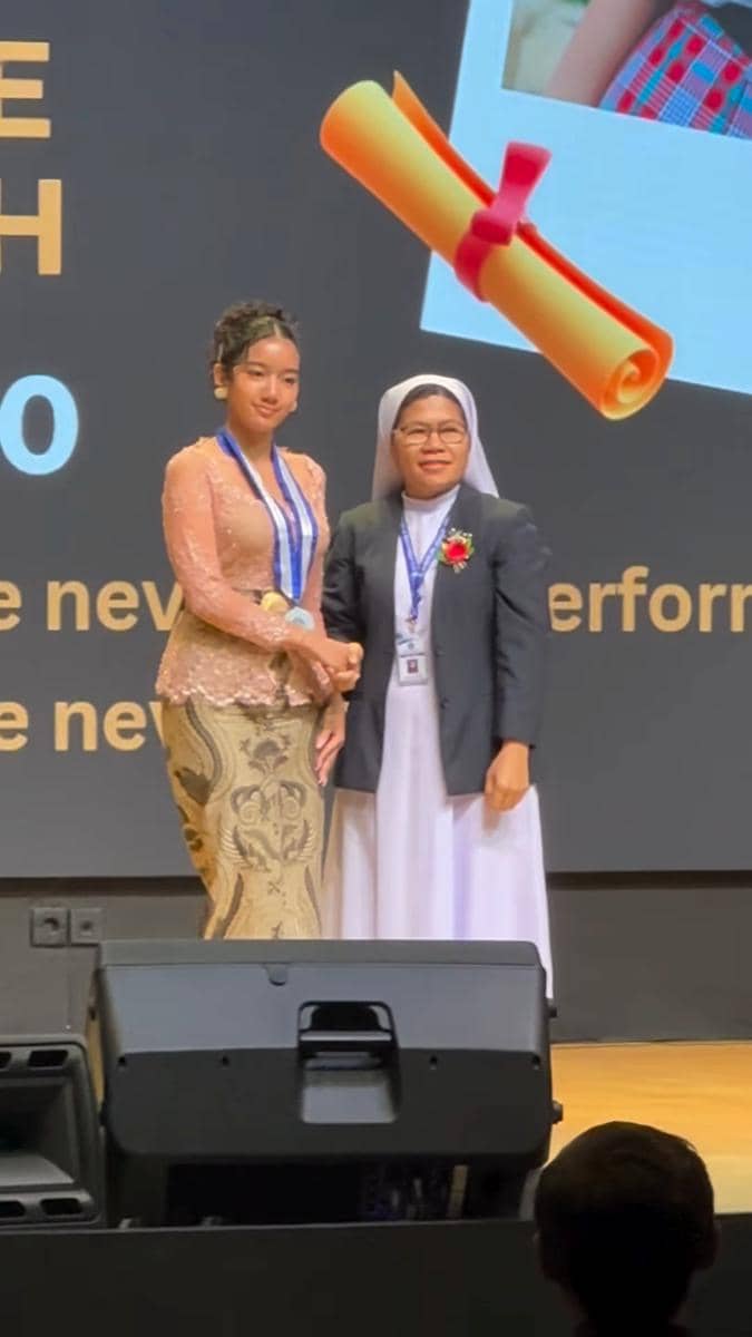 Seorang siswi mengenakan kebaya berpose bersama seorang suster di atas panggung saat acara wisuda SMP.