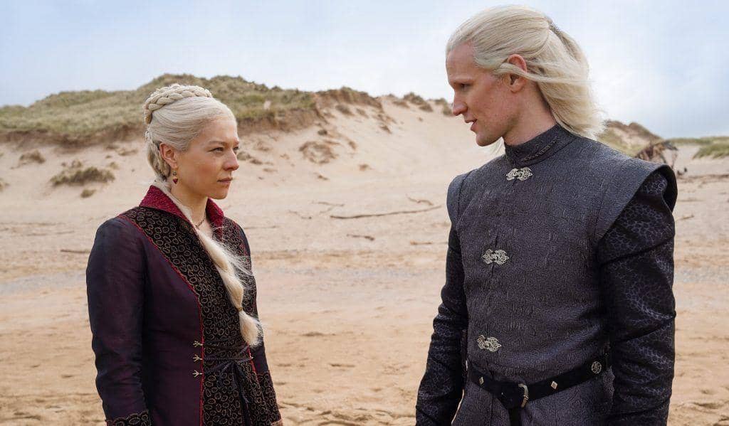 Dua karakter berambut pirang berpakaian bangsawan berdiri di pantai berpasir dalam adegan serial House of the Dragon.