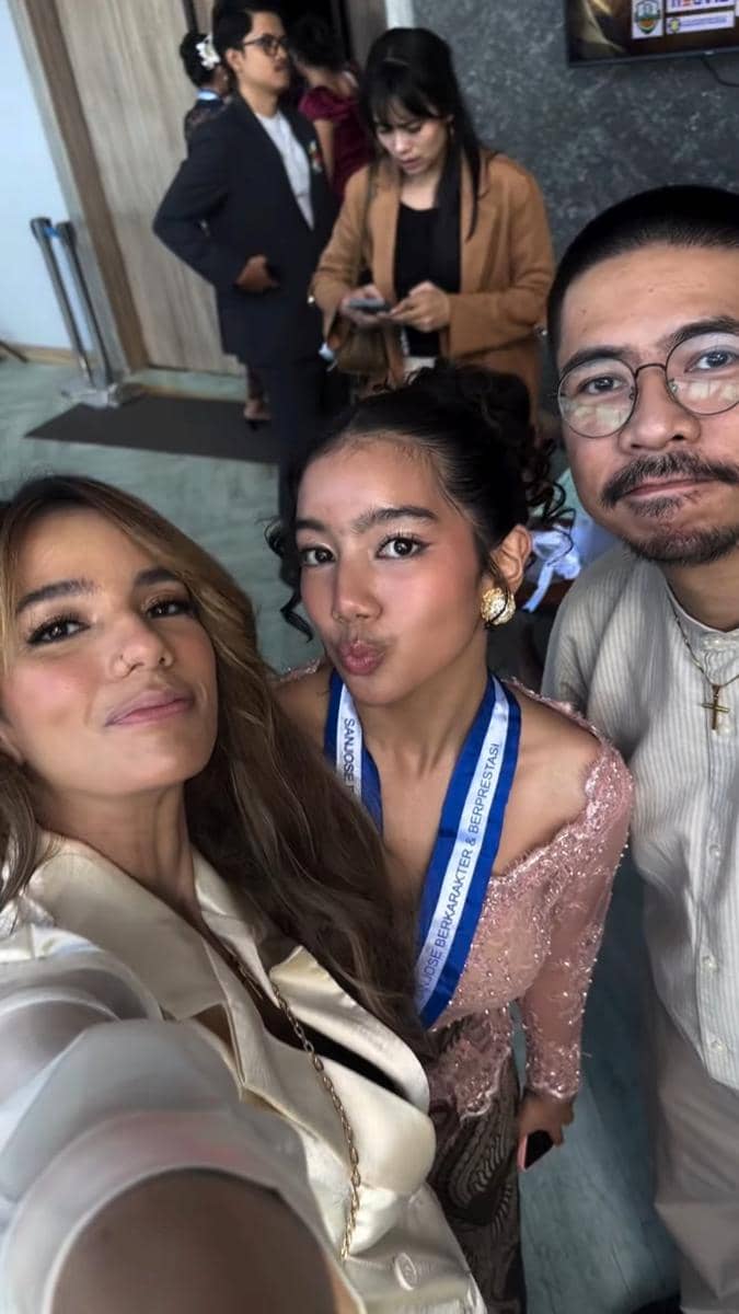 Seorang remaja mengenakan kebaya berwarna pink dengan medali wisuda berpose bersama dua orang dewasa di acara kelulusan SMP.