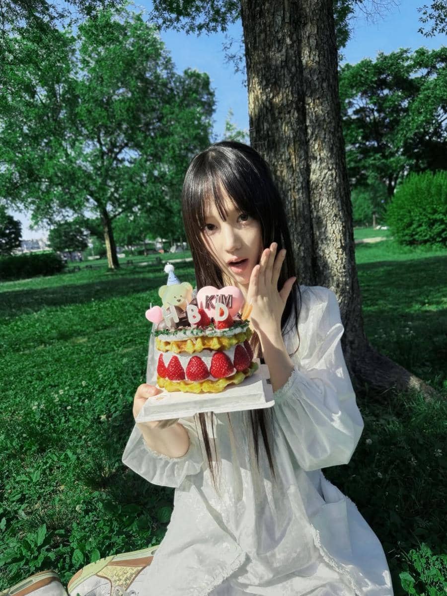 Seorang perempuan berambut panjang mengenakan gaun putih memegang kue ulang tahun bertopping stroberi di taman yang rindang.